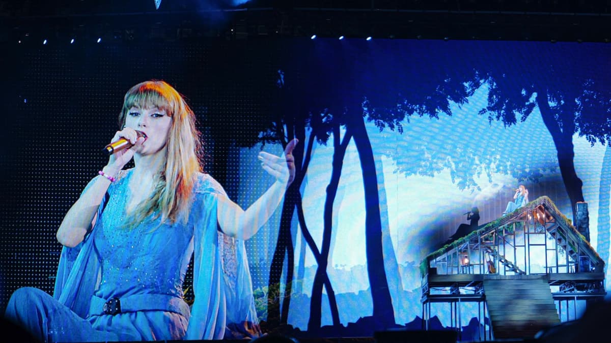 Taylor Swift sitting on a stage rooftop performing a conert in a blue dress