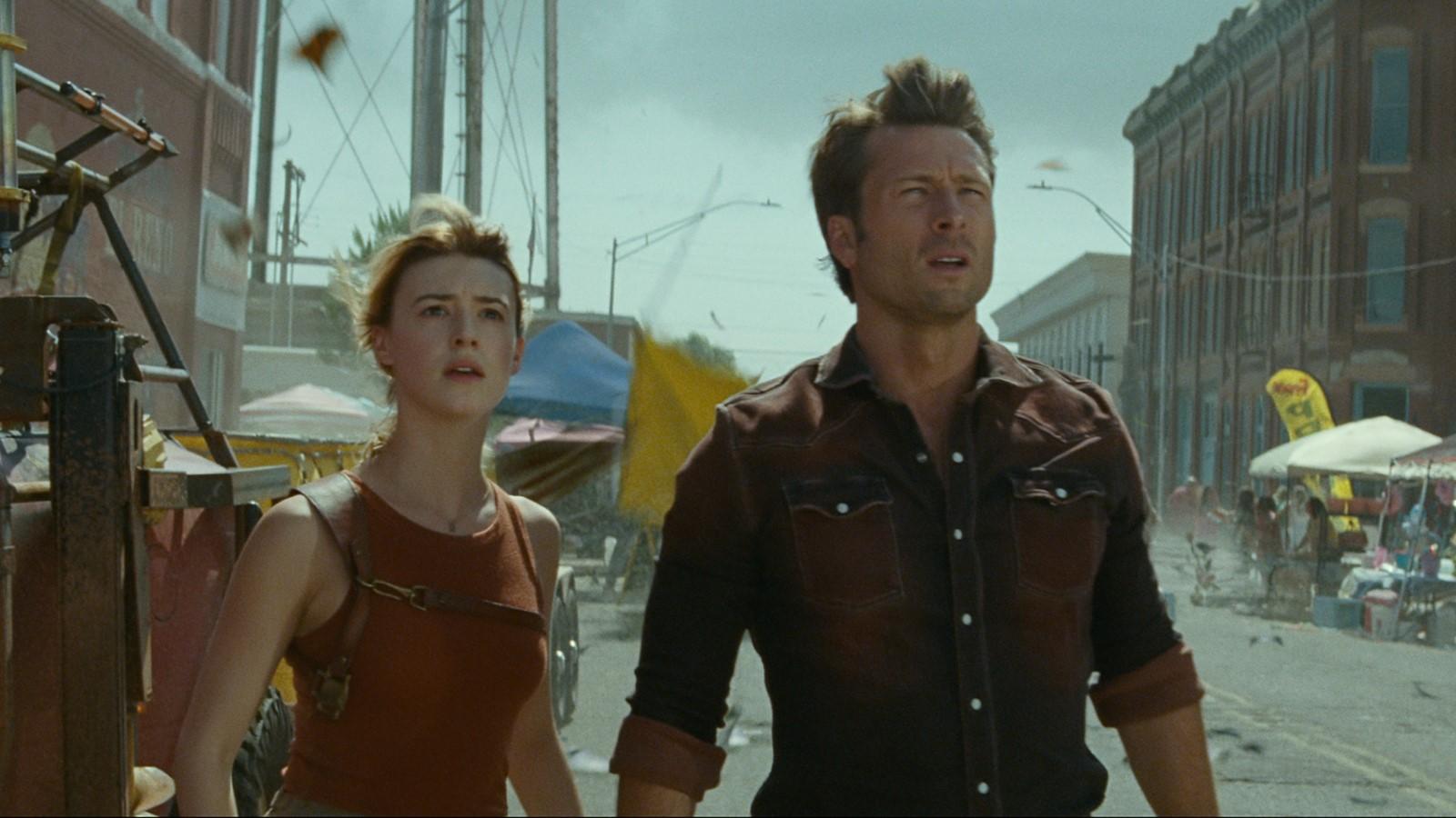 Twisters review: Glen Powell and Daisy Edgar-Jones as Tyler and Kate, standing in the middle of a street and looking past the camera as debris flies past them in Twisters