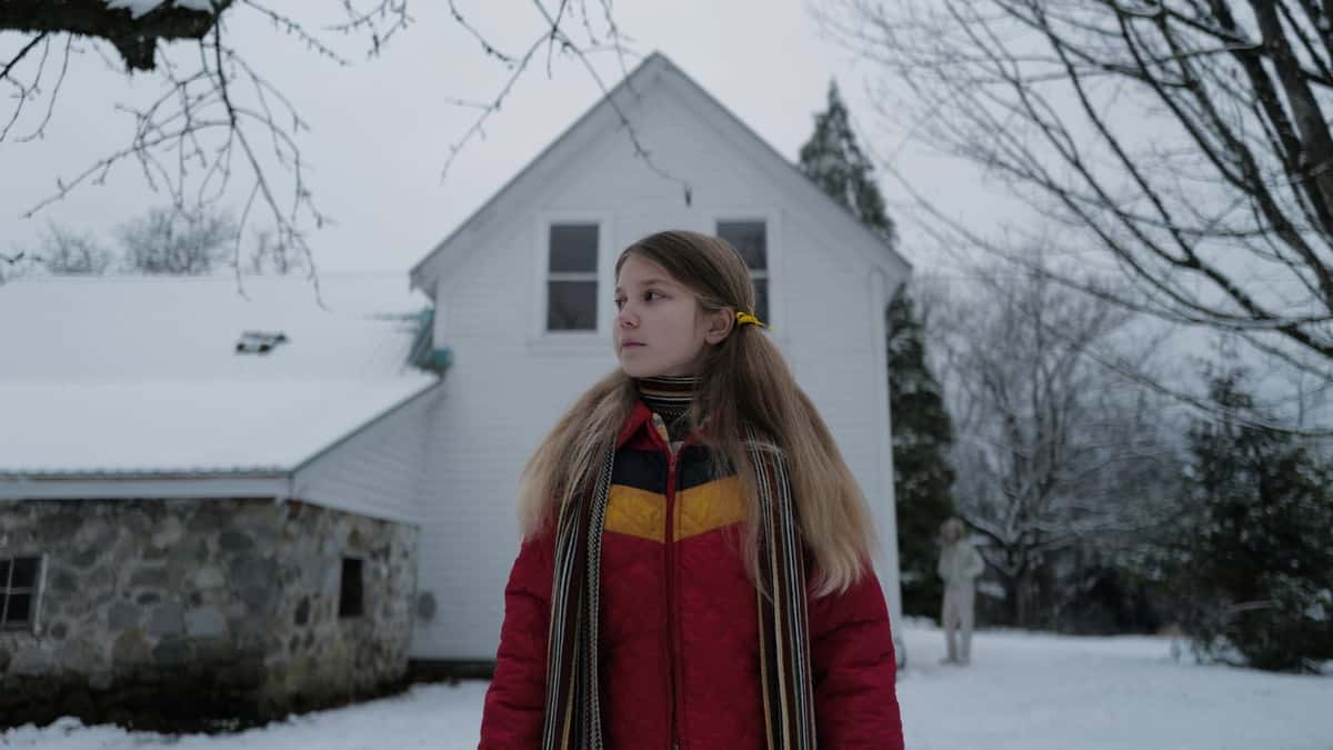 A little girl wearing a red coat standing in front of a white house in the snow in Longlegs