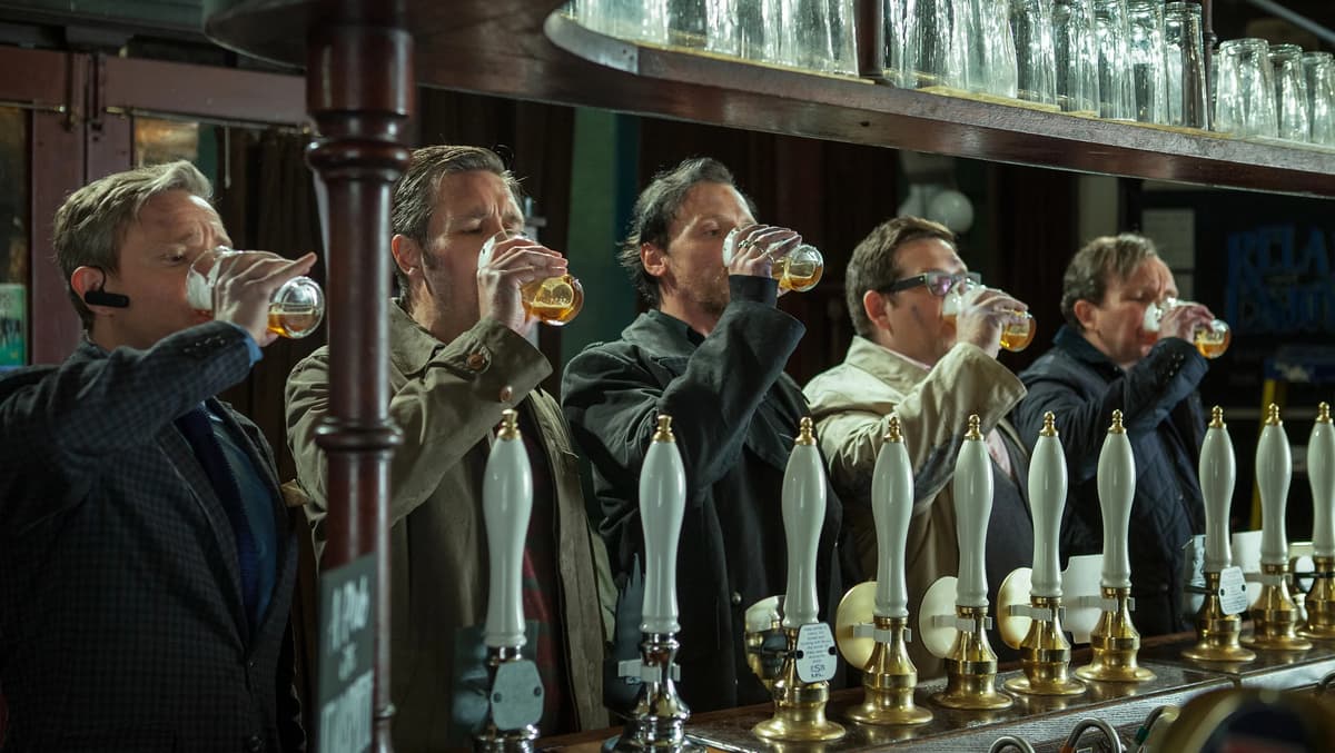 A group of men stand at a bar in a pub and all drink a pint of beer at the same time