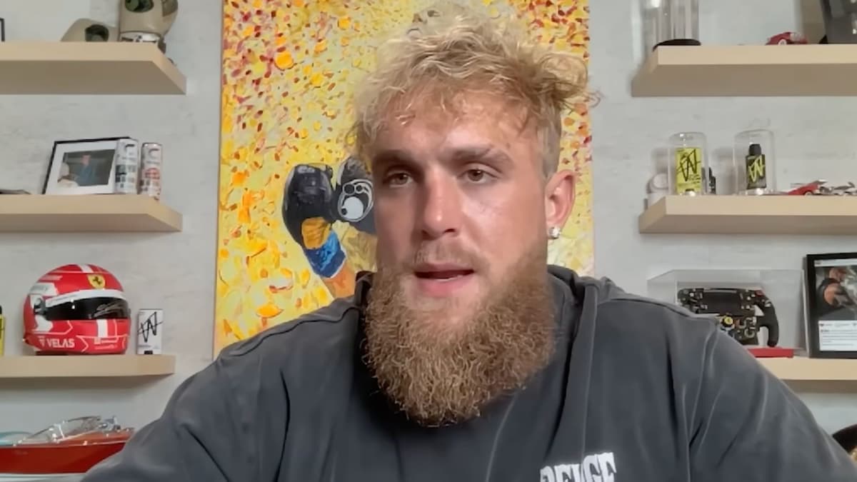 Jake Paul speaking to camera in front of yellow painting and boxing belt