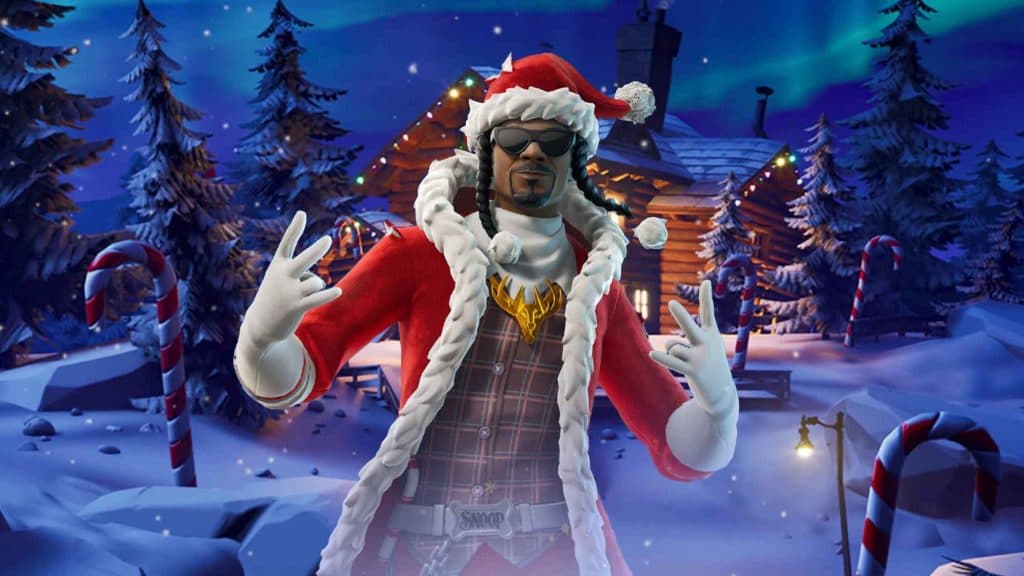 Santa Snoop Dogg, wearing a Santa hat and jacket, posing in front of a log cabin.