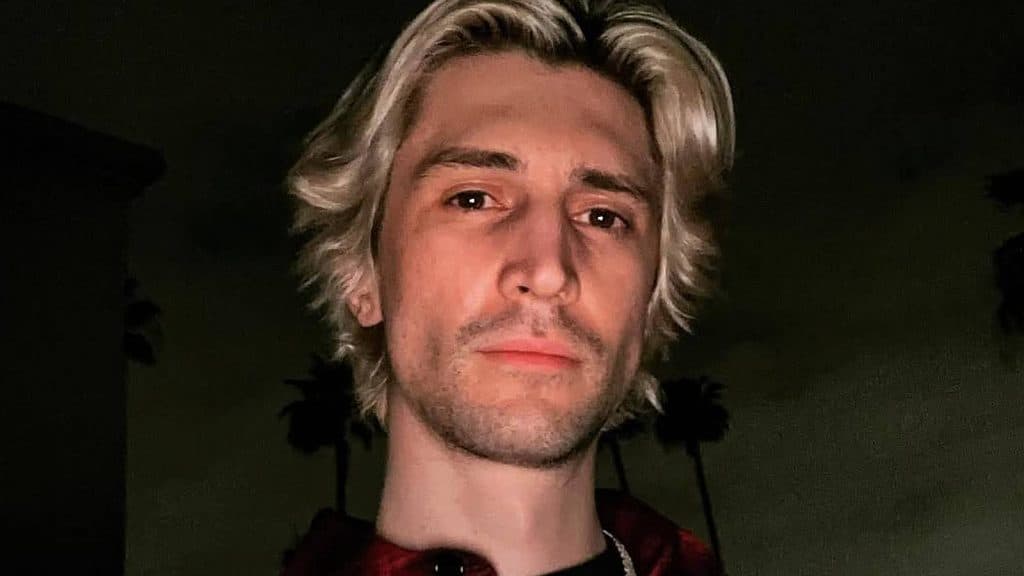 xQc poses for a selfie at night against a sky with palm trees.